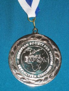 Eine silberne Medaille, in der Mitte das Logo ESPCH und die Umrisse von Europa und Asien, rundherum die Schrift: European Society for Person Centered Healthcare